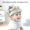 פעוט חובש כובע מגן ראש מרופד עם הדפסים צבעוניים, מחייך בישיבה בתוך הבית. הכובע כולל רצועת סנטר, והילד עונד גם סינר תואם.