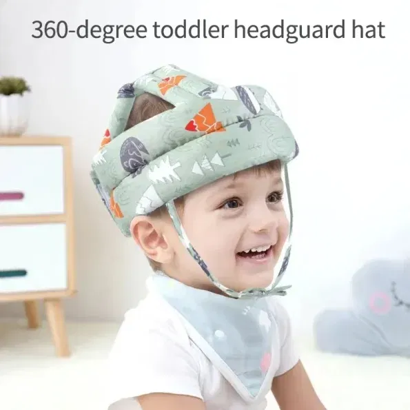 פעוט חובש כובע מגן ראש מרופד עם הדפסים צבעוניים, מחייך בישיבה בתוך הבית. הכובע כולל רצועת סנטר, והילד עונד גם סינר תואם.