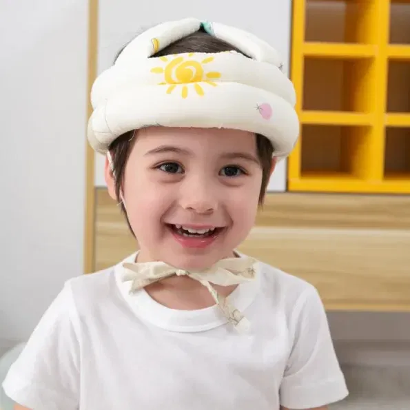 ילד חייכן עונד סרט מגן עם עיצוב שמש, עומד בתוך הבית. הילד לבוש בחולצה לבנה ונראה מאושר. ברקע ארון עץ עם מדפים צהובים.