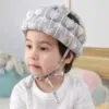 ילד חובש קסדת מגן מרופדת ומעוצבת וחולצת טריקו לבנה יושב בתוך הבית. צעצועים רכים וכריות מטושטשים ברקע. לקסדה בסיס אפור עם עיצובים גיאומטריים לבנים.
