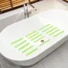 אמבטיה לבנה מודרנית עם פסים ירוקים למניעת החלקה בתחתית. הוא כולל ברז כסף מבריק וממוקם בחדר רחצה עם תריסי עץ הנראים חלקית. הרצפה מרוצפת בגוון אפור בהיר.